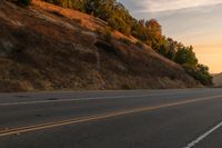 two people ride down the road on a motorcycle at sunset, passing by trees and hills