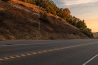 two people ride down the road on a motorcycle at sunset, passing by trees and hills