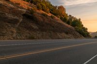 two people ride down the road on a motorcycle at sunset, passing by trees and hills