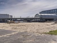 two people walk across the street past buildings and a bridge in front of them and some dirt