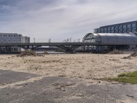 two people walk across the street past buildings and a bridge in front of them and some dirt