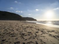 two people walking down a sandy beach at sunset in front of the ocean and hills