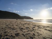 two people walking down a sandy beach at sunset in front of the ocean and hills