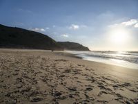 two people walking down a sandy beach at sunset in front of the ocean and hills