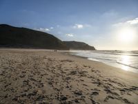 two people walking down a sandy beach at sunset in front of the ocean and hills