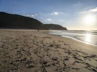 two people walking down a sandy beach at sunset in front of the ocean and hills