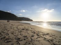 two people walking down a sandy beach at sunset in front of the ocean and hills