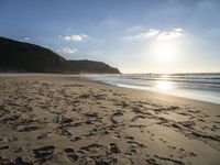 two people walking down a sandy beach at sunset in front of the ocean and hills