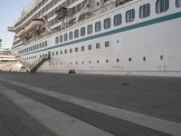 two people walk down an empty sidewalk by a large cruise ship, with stairs to the back and the front