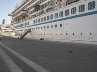 two people walk down an empty sidewalk by a large cruise ship, with stairs to the back and the front