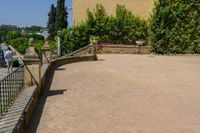 two people walk next to a fence on a path with buildings in the background and trees growing next to the walkway