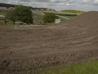 a man on a dirt motocross bike coming in a curve hill, with a green field and farm behind him