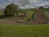 two people on dirt bikes going around a hill course, with green grass, signs and trees behind the track