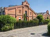 some old buildings and bushes and a brick sidewalk with a bench by the curb and railing