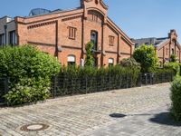 some old buildings and bushes and a brick sidewalk with a bench by the curb and railing