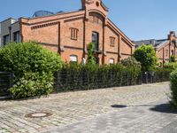 some old buildings and bushes and a brick sidewalk with a bench by the curb and railing