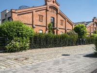 some old buildings and bushes and a brick sidewalk with a bench by the curb and railing