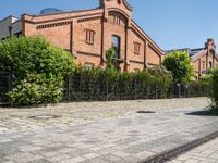 some old buildings and bushes and a brick sidewalk with a bench by the curb and railing