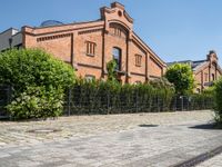 some old buildings and bushes and a brick sidewalk with a bench by the curb and railing