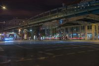 night time shot of city street intersection with urban buildings and elevated bridge over street crossing at night