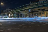 night time shot of city street intersection with urban buildings and elevated bridge over street crossing at night