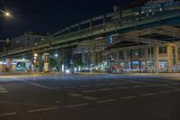 night time shot of city street intersection with urban buildings and elevated bridge over street crossing at night