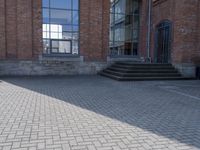 the street is paved with concrete blocks next to a brick building, with stairs and glass windows