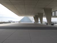 a sidewalk outside a large building with a large glass pyramid behind it by the road