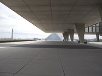 a sidewalk outside a large building with a large glass pyramid behind it by the road