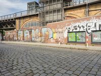 a brick building with graffiti and windows near the sidewalk and stairs and a bridge over it