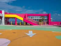 a baseball field in front of a pink building with yellow stairs and a sign pointing to the first