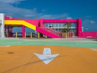 a baseball field in front of a pink building with yellow stairs and a sign pointing to the first