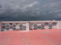 a pink, yellow, and white tennis court under clouds and a sky background and some people on a bench