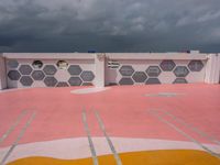 a pink, yellow, and white tennis court under clouds and a sky background and some people on a bench