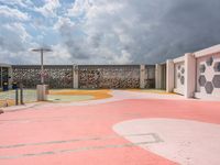 a pink basketball court and a concrete wall with white designs against a cloudy background with a street light