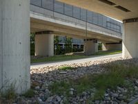 a sidewalk with a bench sitting underneath a bridge over a river area, next to a lawn and the street