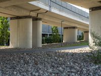 a sidewalk with a bench sitting underneath a bridge over a river area, next to a lawn and the street