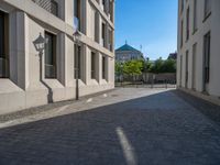 two people walking down an empty city street in an alley area with tall buildings in the background