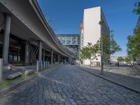 a cobble - stone road in front of an elevated bridge and building on the left, there is no traffic on the side