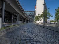 a cobble - stone road in front of an elevated bridge and building on the left, there is no traffic on the side