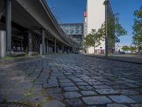 a cobble - stone road in front of an elevated bridge and building on the left, there is no traffic on the side