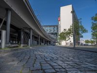 a cobble - stone road in front of an elevated bridge and building on the left, there is no traffic on the side