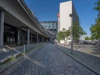 a cobble - stone road in front of an elevated bridge and building on the left, there is no traffic on the side