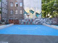 a small basketball court in an urban neighborhood with a graffiti wall behind it and two basketball hoops with basketball balls