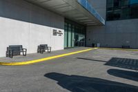 two benches near a large building with a shadow on the sidewalk outside it's entrance