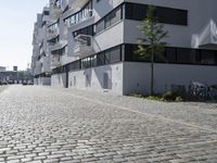 the bricked sidewalk in front of an apartment block in an urban area that contains an old white building with black windows and a white balcony