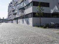 the bricked sidewalk in front of an apartment block in an urban area that contains an old white building with black windows and a white balcony