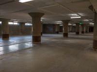 empty parking garage with large pillars and stone floors near light from windows with green exit signs