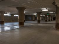 empty parking garage with large pillars and stone floors near light from windows with green exit signs