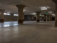 empty parking garage with large pillars and stone floors near light from windows with green exit signs